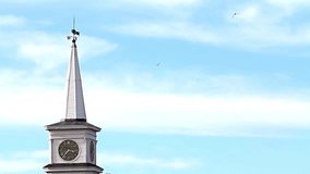 Video of an old New England church clock tower and spire against a blue cloudy sky with birds flying.