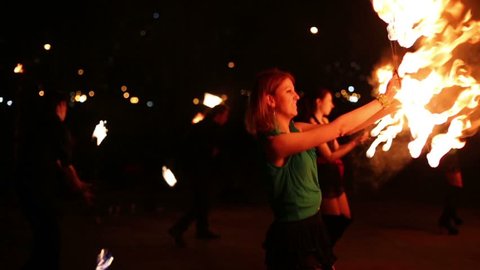 Three girls dance with flaming torches made in shape of fans at evening fire show.
