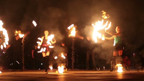 Two girls move with burning fans and four guys rotate pois, staff at evening fire show.
