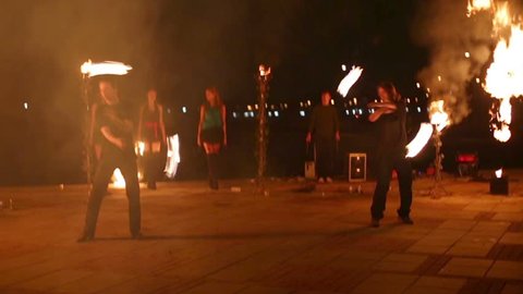 Two boys synchronously move spinning burning pois, three girls stand behind them with fans during evening fire show.

