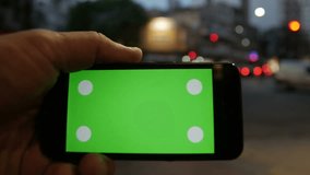 Smart Phone held by hands on the street at dusk, magic hour night. Green screen Chroma Key. Close up. Tracking motion. Horizontal. - 5 s black. 4k