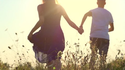 Happy couple having fun outdoors. Couple running away on the meadow. Countryside. Young Man and woman holding hands and running through a field with wild flowers. Sun flare. Slow motion video footage