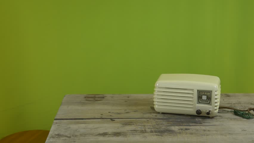 An untidy bizarre man, wearing big patched glasses and a toupee, trying to tune in a station on an old tube bakelite radio, over green background