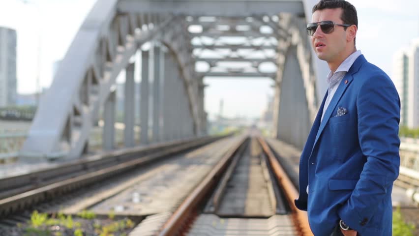 Young man in blue jacket and sunglasses looks at wristwatch on railway bridge