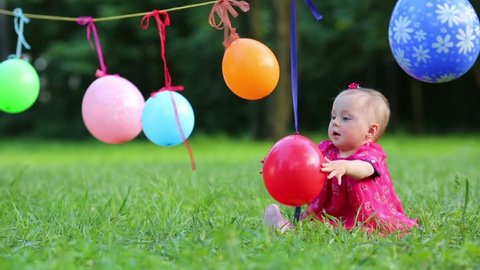 Balloons hang on clothesline and baby in dress catches balloon at summer park