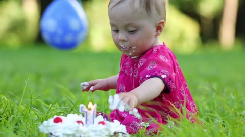 Cute stained baby in dress eats cake with candle in form of 1 at summer park