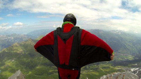 A base jumper in a wingsuit jumps from a cliff, gliding down over a green landscape