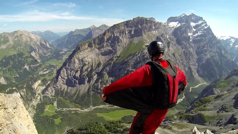 A base jumper in a wingsuit leaping off a from a rocky cliff, gliding down over a green landscape