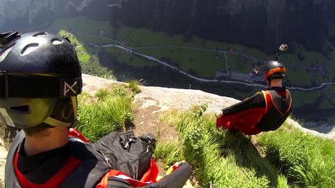 Two base jumpers in wingsuits jumping from a cliff, gliding down over a green landscape, POV