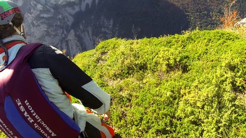 A base jumper runs before leaping off from a cliff and gliding down over a mountain landscape, POV