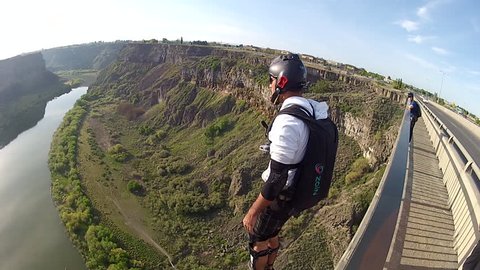 A base jumper jumps down before opening the parachute in mid air, a river visible below