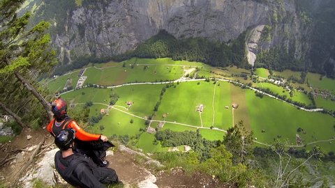 Two base jumpers jumping from a cliff in wingsuits, gliding over a green landscape, POV, top angle
