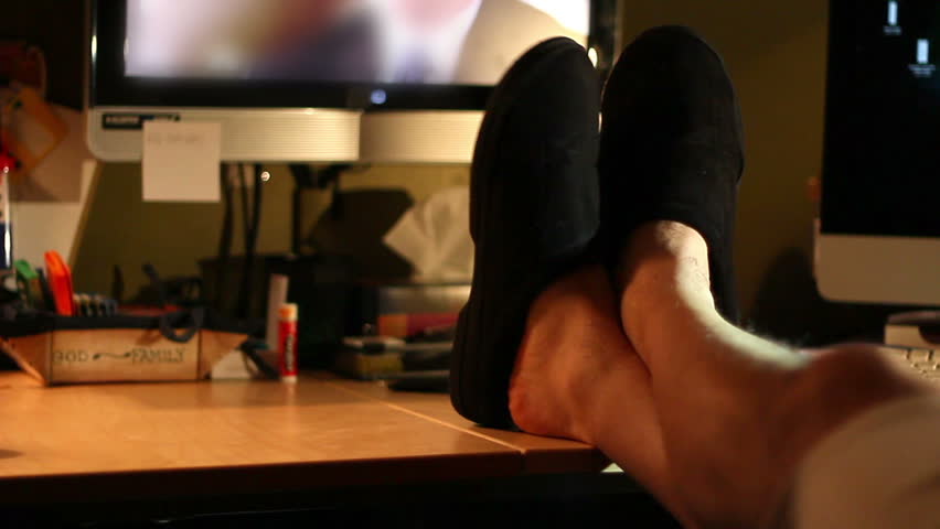 A man in slippers casually watches television with his feet up on his desk.