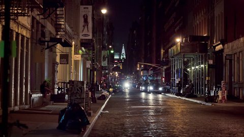 NEW YORK - AUG 1, 2014: people crossing dark SoHo cobblestone street at night with Chrysler Building and car headlights in background. SoHo is a neighborhood south of Houston Street in Manhattan, NYC.