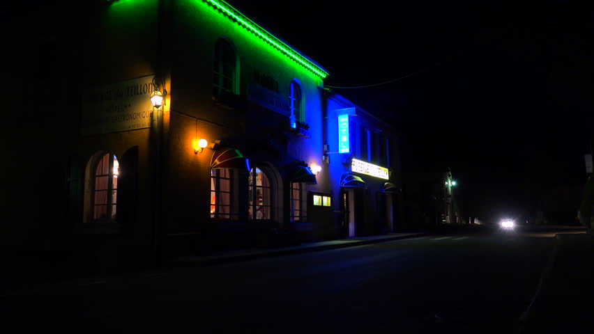 A car passes a small French hotel at night with neon sign flashing.