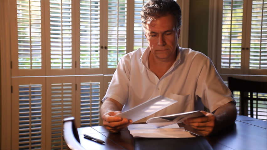 A man sitting at a table in a sunny room looks concerned over the mail he is going through.