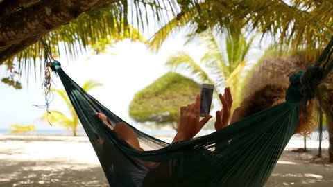 Young Woman with Curly Hair Using Mobile Smart Phone while Relaxing in a Hammock on the Beach. Exotic Island Koh Phangan. Thailand. HD, 1920x1080.