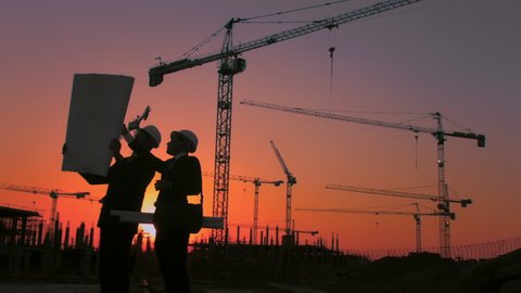 Steadicam shot of skyline and cranes with Asian construction/ executive/ architect/ engineer consultant discussing blue print of new urban development with caucasian construction executive at sunset.