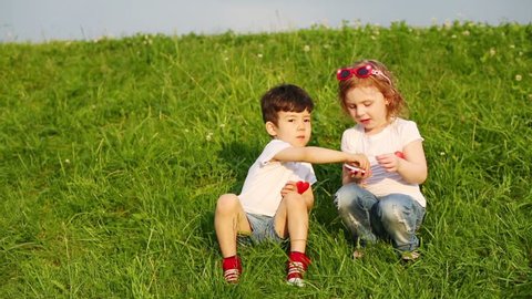 Two children with red hearts on shirts sit on green grass and eat candy