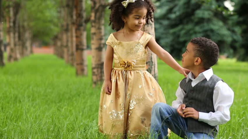 Little girl in beautiful gown stands and boy squats near she in park and show on the fingers of her age