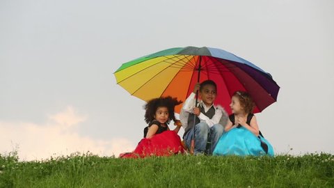 Three children sit on grass under colorful umbrella during rain and laugh