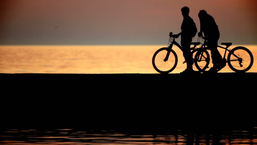 Silhouettes of two cyclists at sunset in the sea. They go on pier