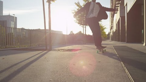 SLOW MOTION: Cool young businessman longboarding to work. Cheerful businessman with arms raised celebrating while skating along the downtown street. Ecstatic businessman skateboarding at golden sunset
