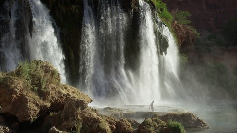 Wide slow motion shot of man walking in rocky waterfall / Havasupai, Arizona, United States