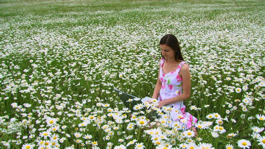 A girl sits on a field with a computer. Young beautiful girl sitting on chamomile field and working on the laptop. Then throws her computer and placed on chamomile field.