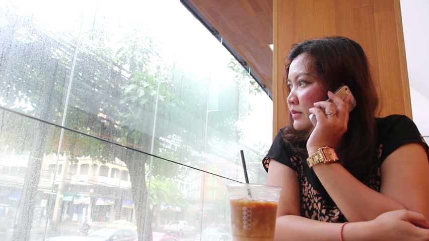 Closeup to Asian girl talking on mobile phone in coffee shop, drinking coffee, raining outside on street