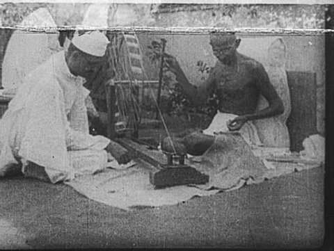 INDIA- CIRCA LATE 1920's: Gandhi spins yarn, assisted by a man in a topi and mundu. Children look on, holding yarn in their hands.
