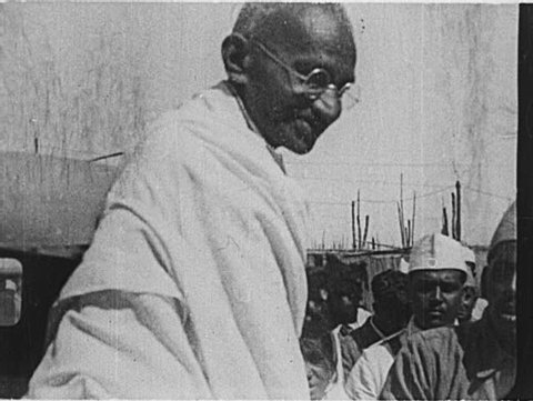 INDIA- CIRCA 1930: A large crowd greets Gandhi as he steps out of a car and acknowledges its members.  Men crowd around him, offering gifts and flowers, praying, and bowing.
