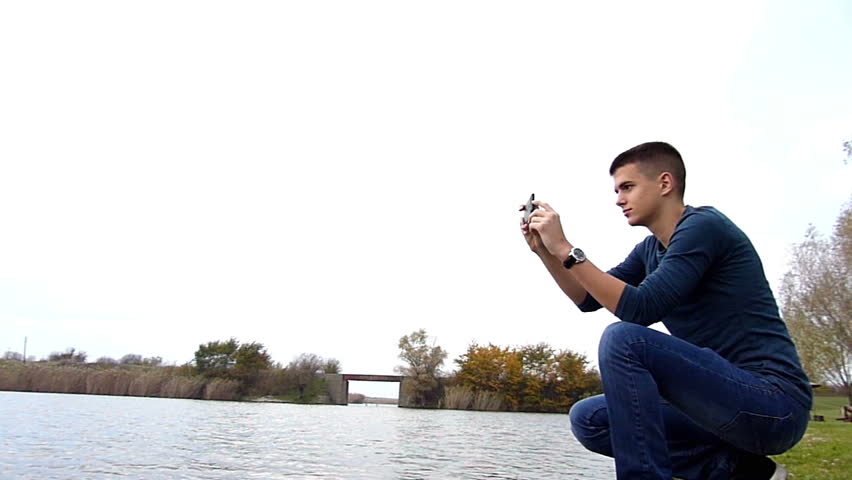 Young man taking photos with smart phone on lake shore with his younger brother. Cloudy fall day. High speed camera,slow motion video,dolly slider.