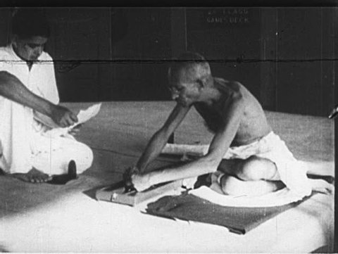 INDIA- CIRCA 1930: Gandhi sits and spins yarn on a loom. Gandhi descends steps, walks through village street, and approaches a home with a young man in a mundu.

