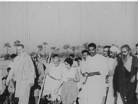 INDIA- CIRCA 1930: A group walks alongside Gandhi.  Gandhi walks a crowd.  He listens to a supporter. He sits. Speaks into a microphone.  He sits on a pillow. Two girls lead him through a crowd.
