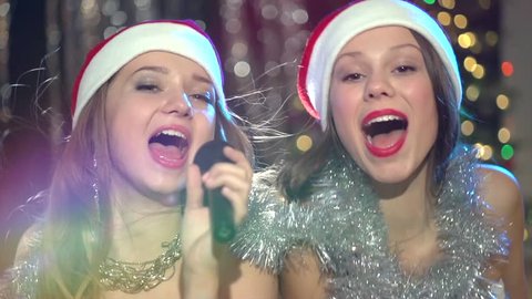 Christmas karaoke party. Two beautiful teenage girl in Santa hat singing over Glowing holiday background. Young cheerful women celebrating Christmas and New Year. HD video footage HD 1080. Slow motion