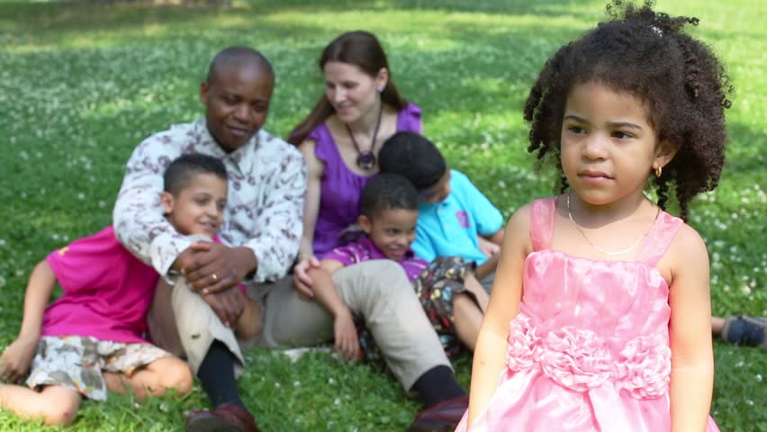 Little girl in pink dress standing in the park, her family sitting on the grass behind | Shutterstock HD Video #7929739