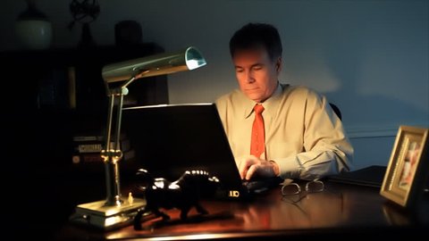 A businessman working late, stops, looks at a framed picture, closes his laptop, turns off the desk lamp, and starts to leave his office.