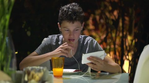 Young teenager eating and watching movie on smartphone at home at night
