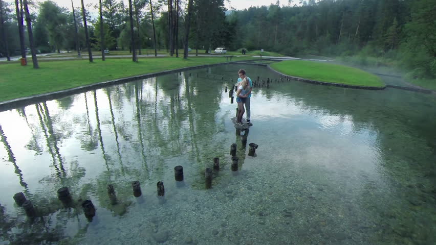 Boy and girl jumping on wooden stump on water. Aerial shot of young romantic happy couple enjoying time on vacation walking on stumps looking out of small lake.