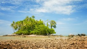 Video - Small tropical island at low tide. under a blue sky with cirus clouds. Lots of coconut palms and other trees. A rocky beach with boulders. Shallow tide-pool in the foreground