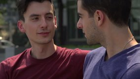 A close-up of two Caucasian gay men, sitting and talking on a bench. Both look to camera.