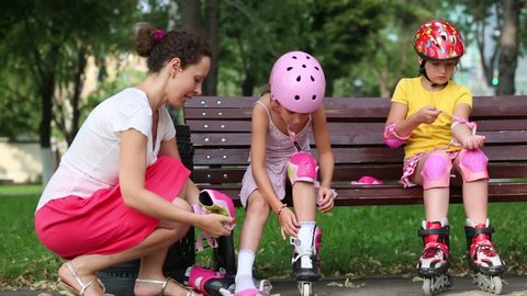 Woman helps two girls put on rollers and protective knee and elbow pads