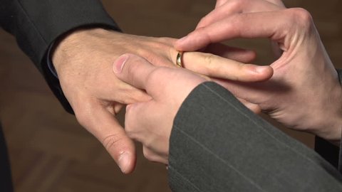 A close-up of the hands of two Caucasian gay men as they put wedding rings on each other.