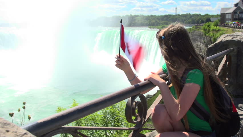 A mid-shot of a young girl on vacation at Niagara Falls in Canada. She turns, looks to camera, smiles and gives a thumbs-up.