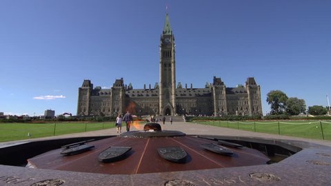 A pan left across the Centennial Flame, in front of Parliament, Ottawa, Canada as tourists walk towards the building.