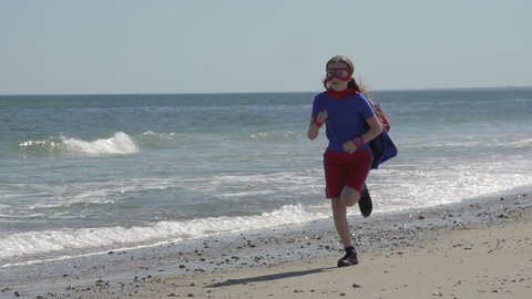 A wide shot of a young girl, dressed as a superhero running down a beach in slow motion, heroically.