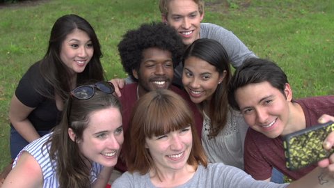 A group of racially-diverse students, gather together as a female student holds up a cell phone to capture a selfie photograph.