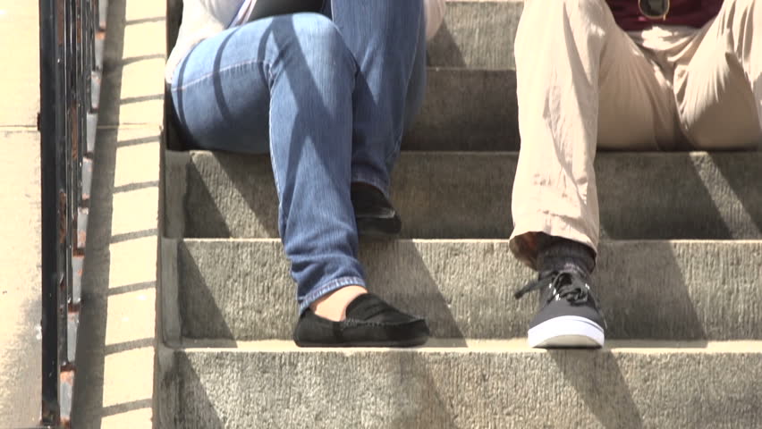 A tilt up to a mid-shot of a black male student and a hispanic female student, clasping class notes. chatting casually on school steps as other students pass.