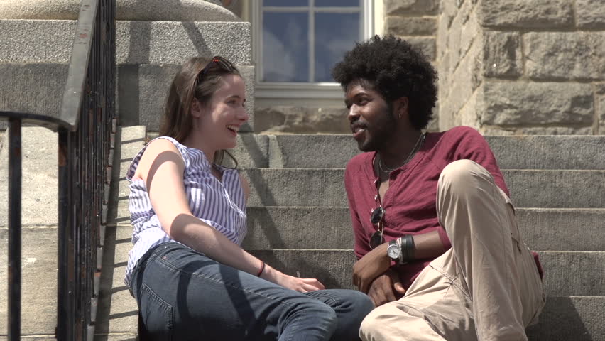 A mid-shot of a black male student and a white female student, sitting on the school steps, talking as other students pass.